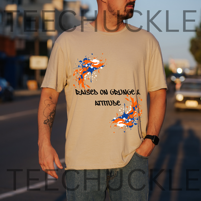 A man wearing a sand graphic t-shirt with orange and blue paint splatters and the text 'RAISED ON GRUNGE & ATTITUDE'. The background is a blurred street at sunset