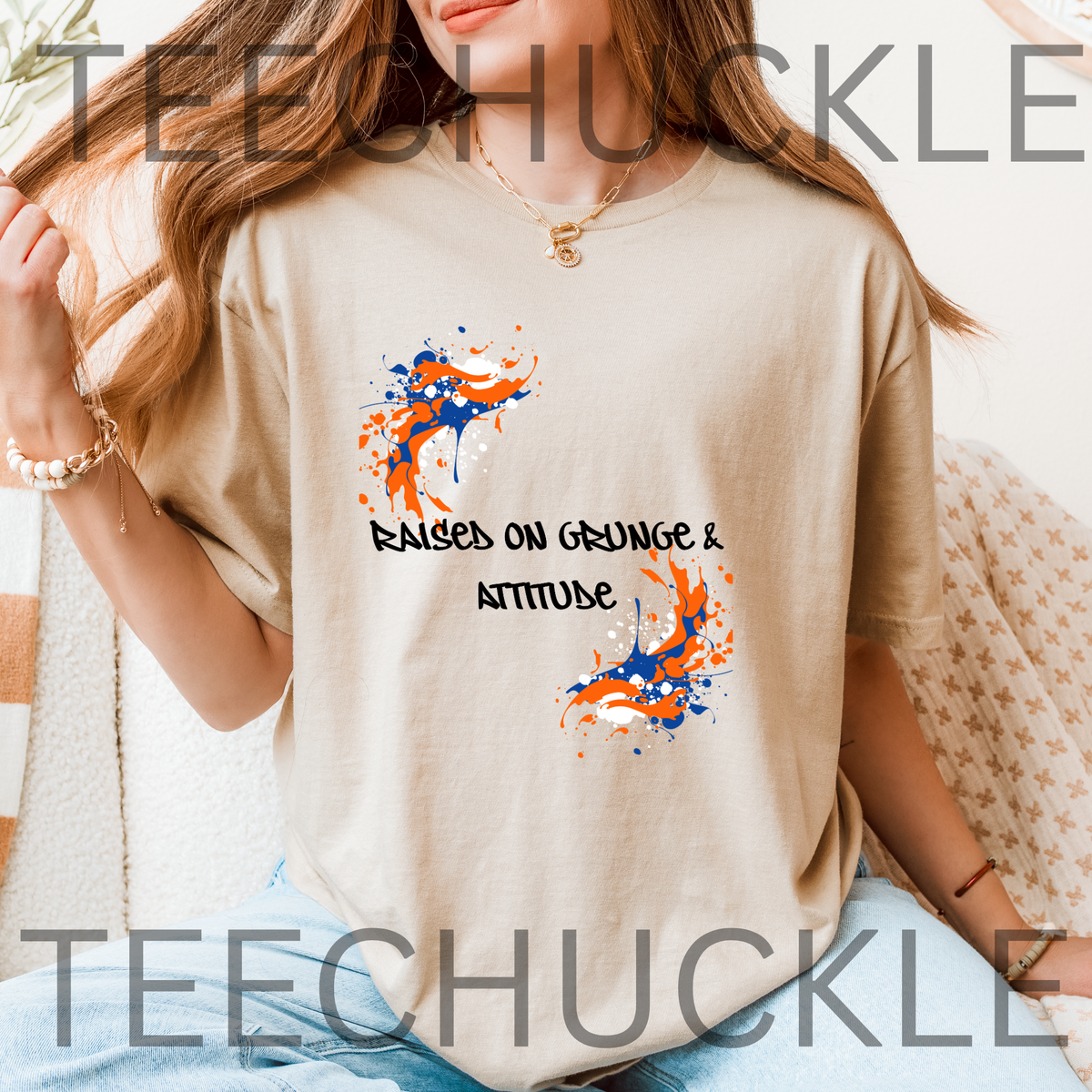 A woman wearing a sand graphic t-shirt with orange and blue paint splatters and the text 'RAISED ON GRUNGE & ATTITUDE'.