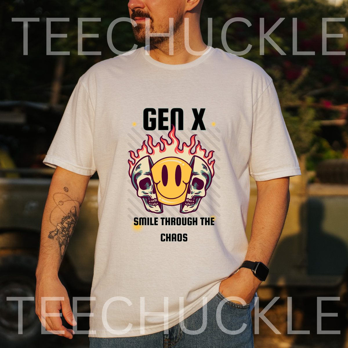 A man wearing a white graphic t-shirt with a 'Gen X Smile Through The Chaos' design featuring skulls, a yellow smiley face, and flames. The photo is taken outdoors in warm, golden-hour lighting