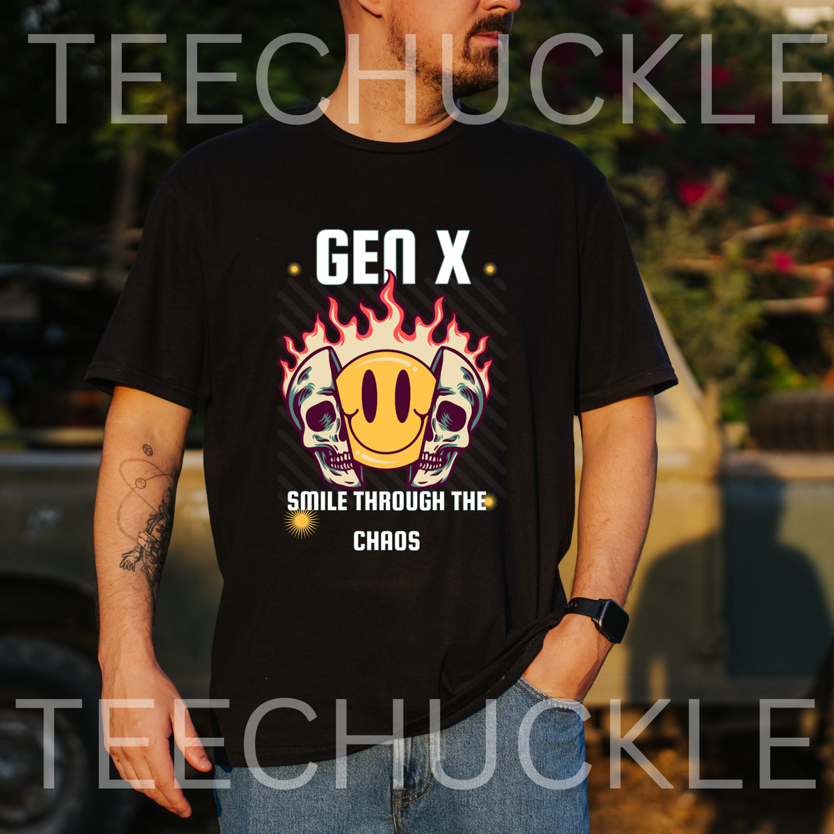 A man wearing a black graphic t-shirt with a 'Gen X Smile Through The Chaos' design featuring skulls, a yellow smiley face, and flames. The background is an outdoor blurred setting