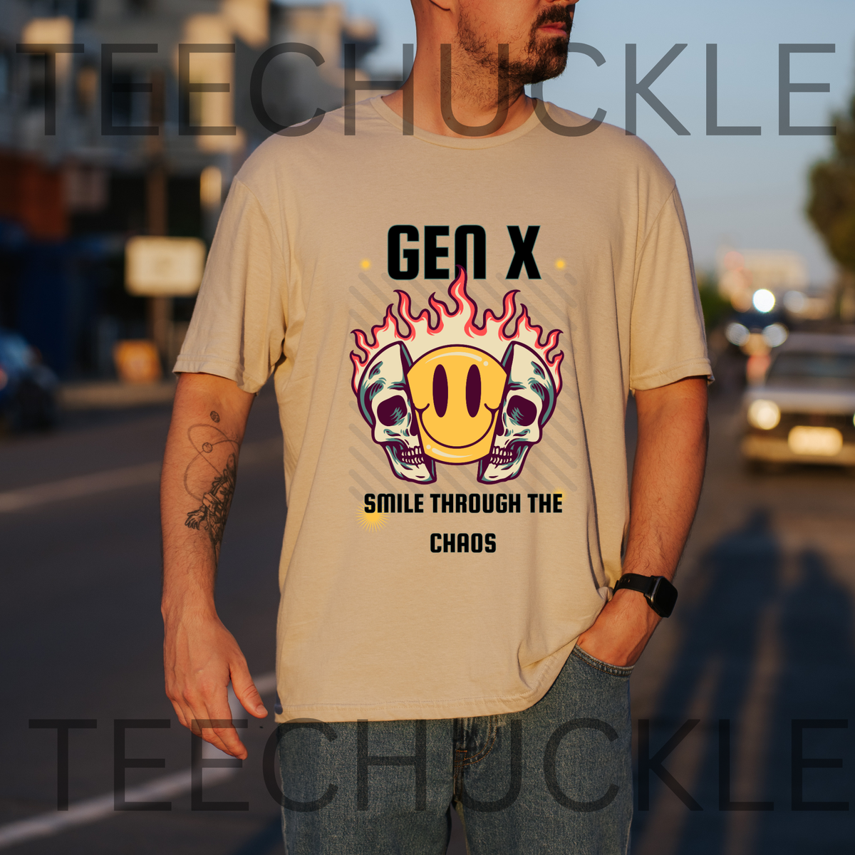 A man wearing a sand graphic t-shirt with a 'Gen X Smile Through The Chaos' design featuring skulls and a smiley face in flames, standing outdoors on a street at sunset.
