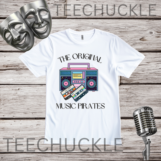 A white graphic t-shirt featuring a retro boombox and cassette tape design with the text 'THE ORIGINAL MUSIC PIRATES'. The t-shirt is displayed on a rustic wood background with two silver theater masks, a vintage microphone