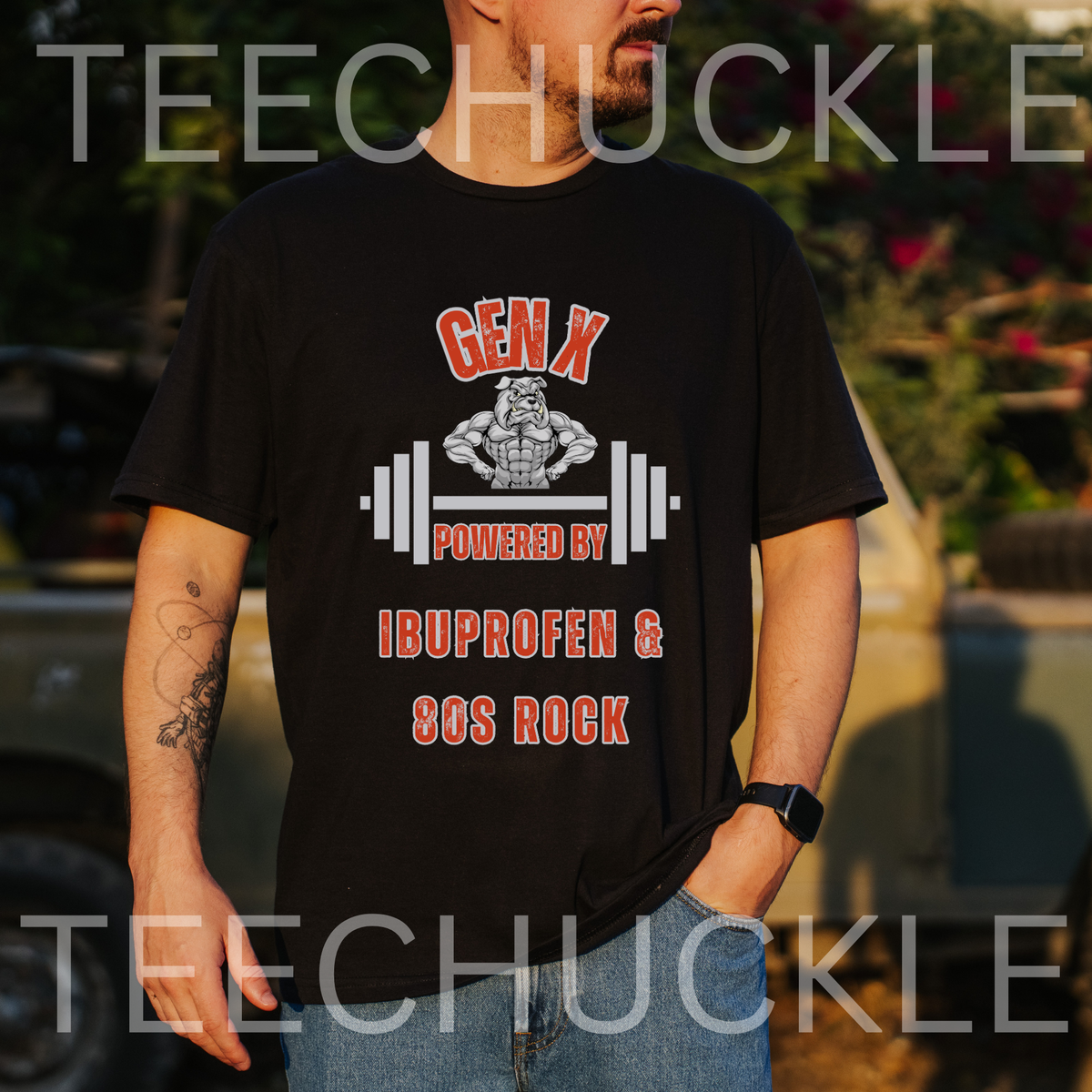 A man wearing a black graphic t-shirt with a red and white design that reads 'GEN X POWERED BY IBUPROFEN & 90S ROCK' with a barbell and cartoon figure. The background is a blurred outdoor setting .