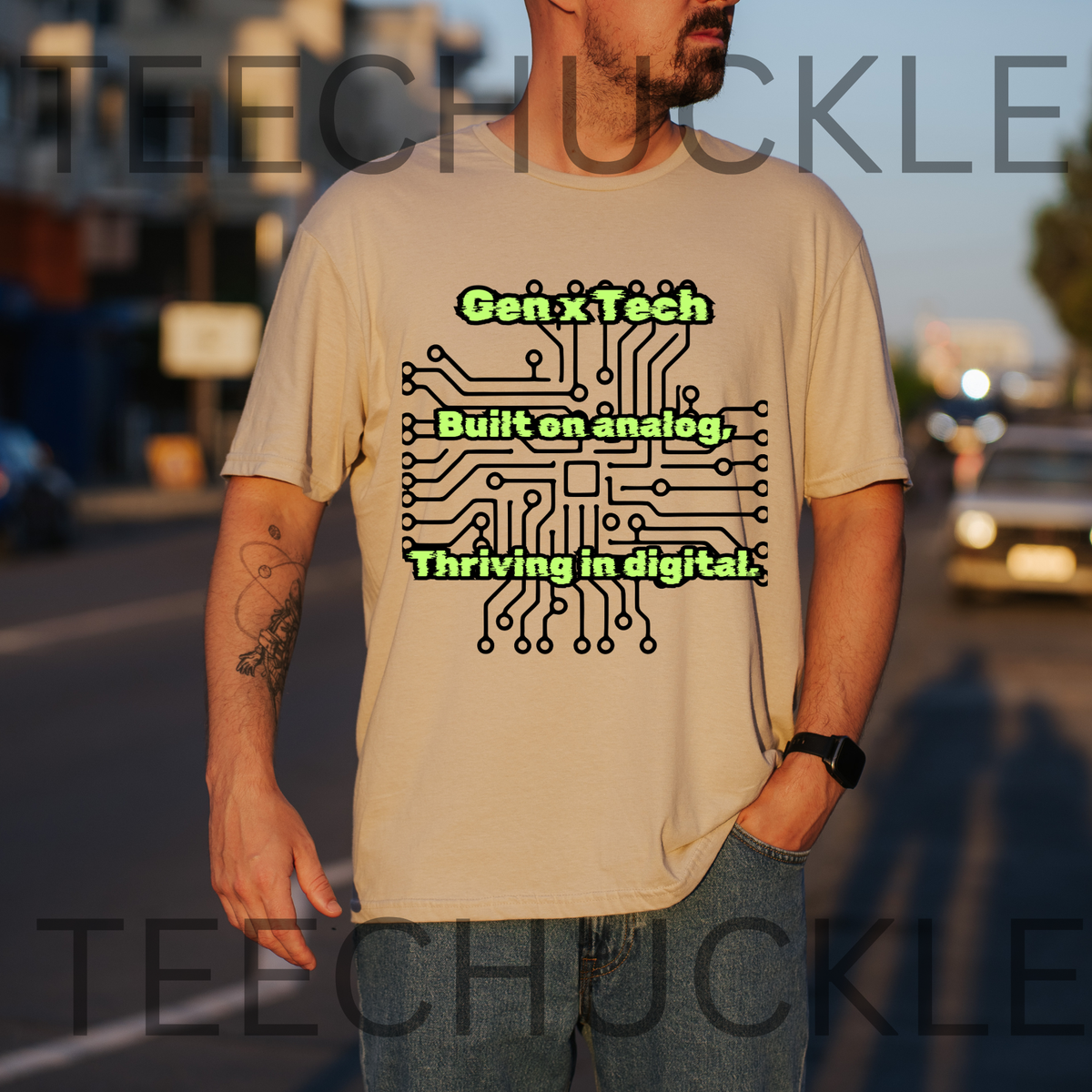A man wearing a sand graphic t-shirt with a black circuit board design and green text 'Gen x Tech Built on analog, Thriving in digital.' standing on a street at sunset.