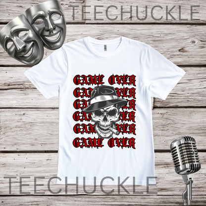 A white short-sleeve graphic T-shirt laid flat on a wood plank background, featuring a skull in a hat smoking a cigerette design with the text 'GAME OVER' repeatedly stacked. Decorative vintage microphones and a microphone and comedy masks appear at the top left and bottom right.