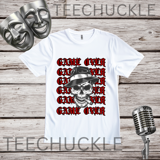 A white short-sleeve graphic T-shirt laid flat on a wood plank background, featuring a skull in a hat smoking a cigerette design with the text 'GAME OVER' repeatedly stacked. Decorative vintage microphones and a microphone and comedy masks appear at the top left and bottom right.