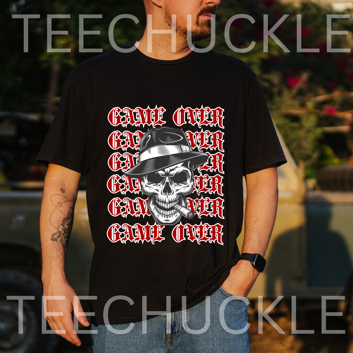 A man wearing a black graphic t-shirt with a repeating 'GAME OVER' text design in red and white around a central skull wearing a hat and smoking a cigar. The background is an outdoor blurred setting with a car.