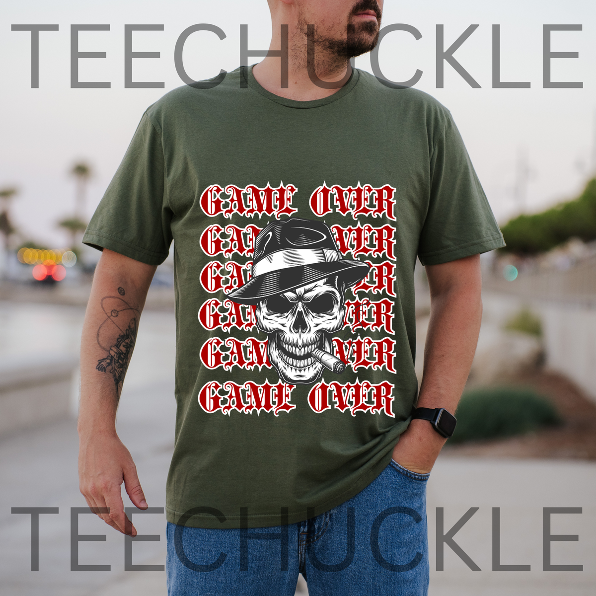 A man wearing a green graphic t-shirt with a repeating red 'GAME OVER' text design and a central skull wearing a hat. The background is a blurred outdoor street scene