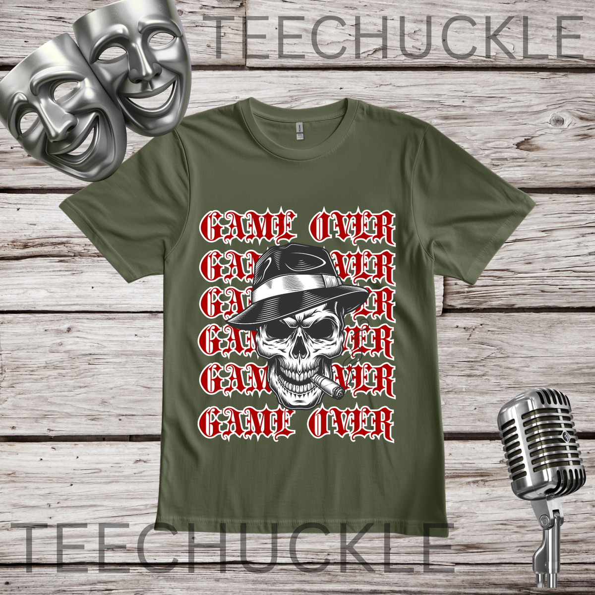 A green graphic t-shirt featuring a repeating 'GAME OVER' text design in red and white, overlaid with a black and white skull wearing a fedora hat. The t-shirt is displayed on a rustic wooden background with silver theater masks and a vintage microphone prop