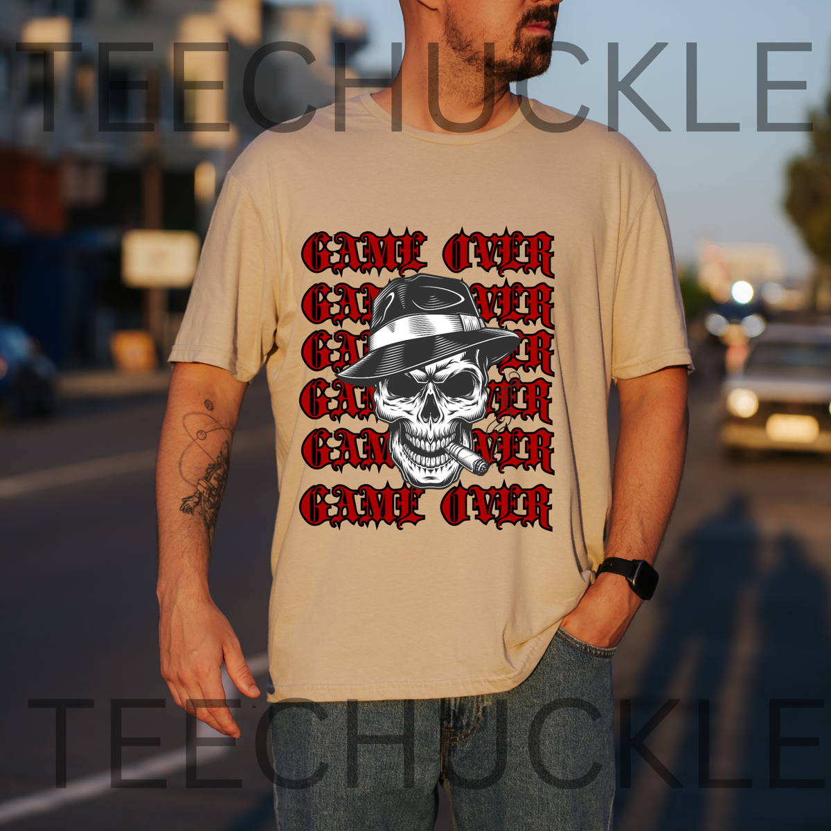 A man wearing a sand graphic t-shirt with a repeating red and black 'GAMEN OVER' design and a skull wearing a hat. The background is a blurred street scene at sunset