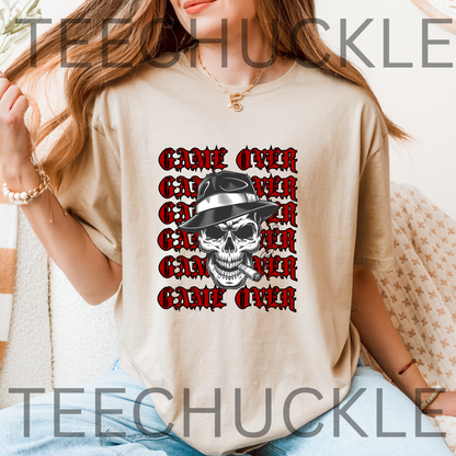 A woman wearing a sand graphic t-shirt with a repeating red and black 'GAME OVER' gothic font design featuring a central black-and-white skull wearing a hat. 