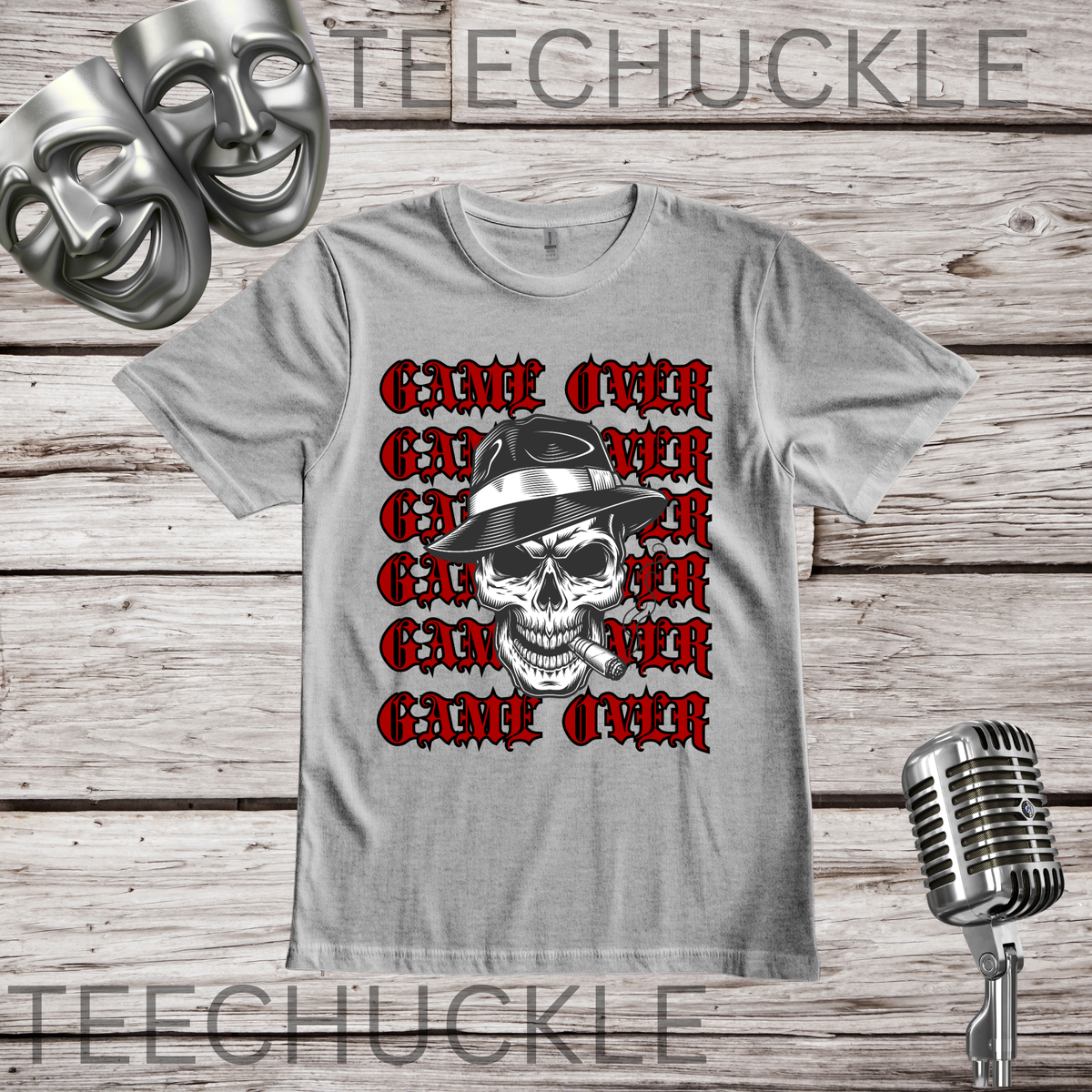 A gray graphic t-shirt featuring a repeating red 'GAME OVER' text design with a central black and white skull wearing a fedora. The shirt is displayed on a rustic wooden background with a vintage microphone and comedy masks