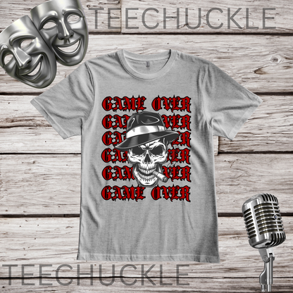 A gray graphic t-shirt featuring a repeating red 'GAME OVER' text design with a central black and white skull wearing a fedora. The shirt is displayed on a rustic wooden background with a vintage microphone and comedy masks