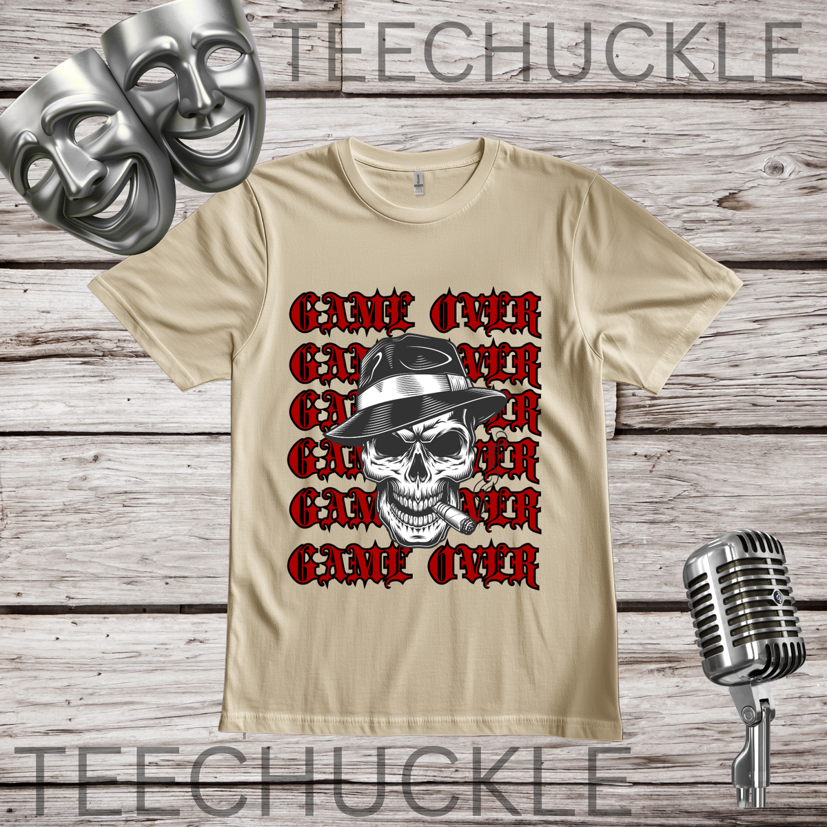 A sand short-sleeve graphic T-shirt laid flat on a wood plank background, featuring a skull in a hat smoking a cigerette design with the text 'GAME OVER' repeatedly stacked. Decorative vintage microphones and a microphone and comedy masks appear at the top left and bottom right.