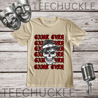 A sand short-sleeve graphic T-shirt laid flat on a wood plank background, featuring a skull in a hat smoking a cigerette design with the text 'GAME OVER' repeatedly stacked. Decorative vintage microphones and a microphone and comedy masks appear at the top left and bottom right.