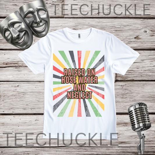 A white graphic t-shirt displayed on a rustic wooden background, featuring a retro sunburst design in red, yellow, green, and gray with the text 'RAISED ON HOSE WATER AND NEGLECT'. Two silver comedy masks are on the left, and a vintage microphone is on the right.