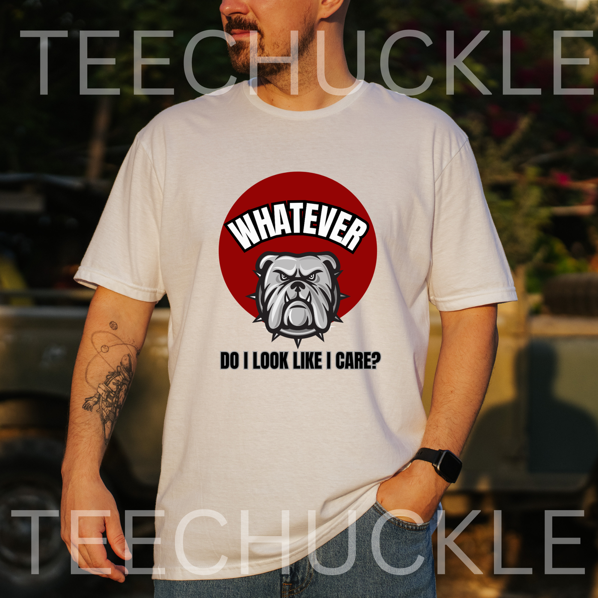 A man wearing a white graphic t-shirt with a red circular bulldog design and the text 'WHATEVER DO I LOOK LIKE I CARE?' on the front, set against a blurred outdoor background. 