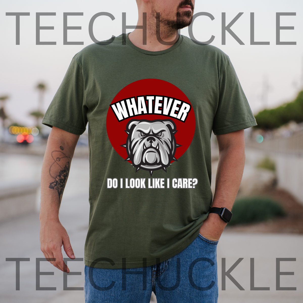 A man wearing a green graphic t-shirt with a red circular bulldog design and the text 'WHATEVER DO I LOOK LIKE I CARE?' on the front, set against an outdoor blurred background. 