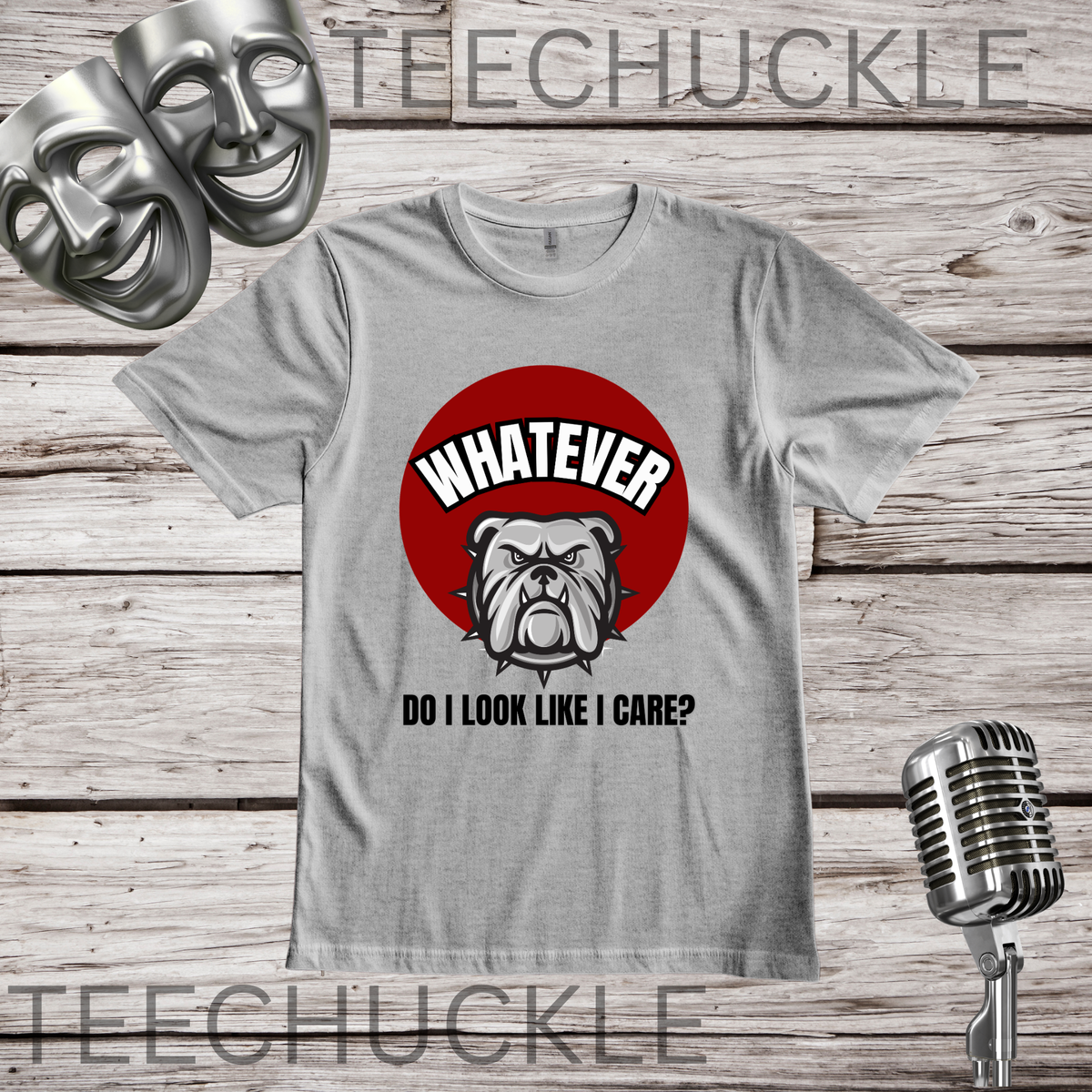 A gray graphic t-shirt featuring a red circle with a bulldog illustration and the text 'WHATEVER DO I LOOK LIKE I CARE?' on a rustic wood plank background, with a vintage microphone and comedy masks as props.