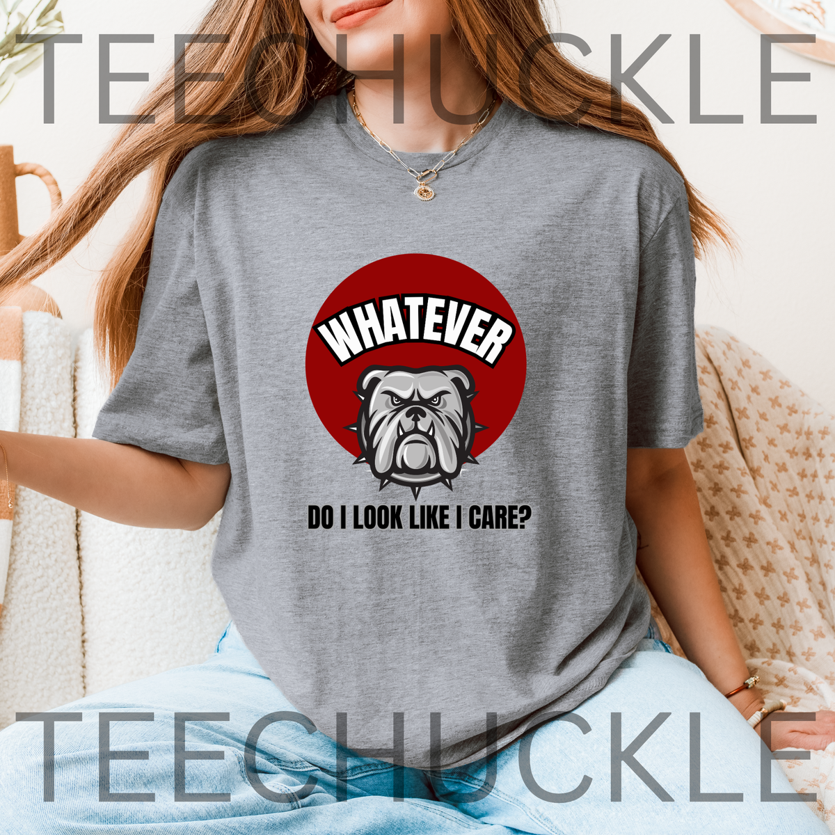 A woman wearing a gray graphic t-shirt with a red circular bulldog design and the text 'WHATEVER DO I LOOK LIKE I CARE?' on the front, seated in a cozy indoor setting.