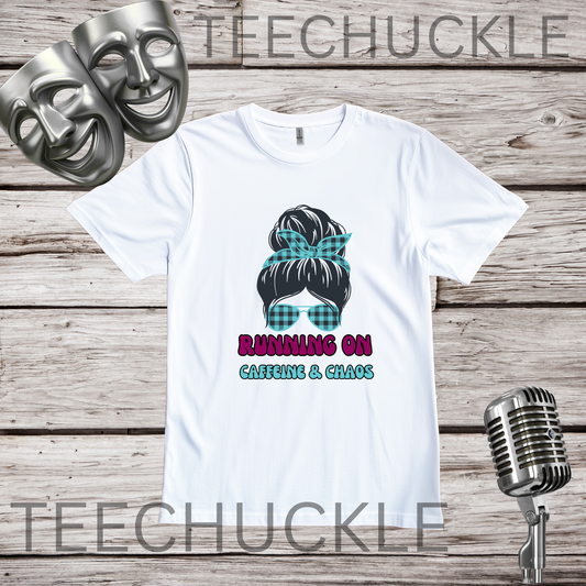 A white graphic t-shirt displayed on a rustic wood background, featuring a design of a woman's hair in a messy bun with a blue plaid bow and heart-shaped sunglasses, with the text 'RUNNING ON CAFFEINE & CHAOS' in pink and blue. Two silver theater masks are on the left, and a vintage microphone is on the right.