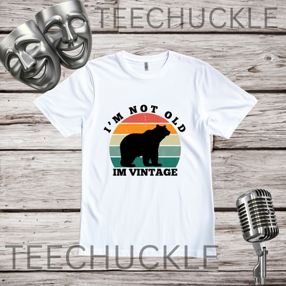 A white short-sleeve graphic T-shirt laid flat on a rustic wood background, with two silver theater masks and a vintage microphone as props. The shirt has a retro sunset-style bear graphic and the text 'I'M NOT OLD I'M VINTAGE.