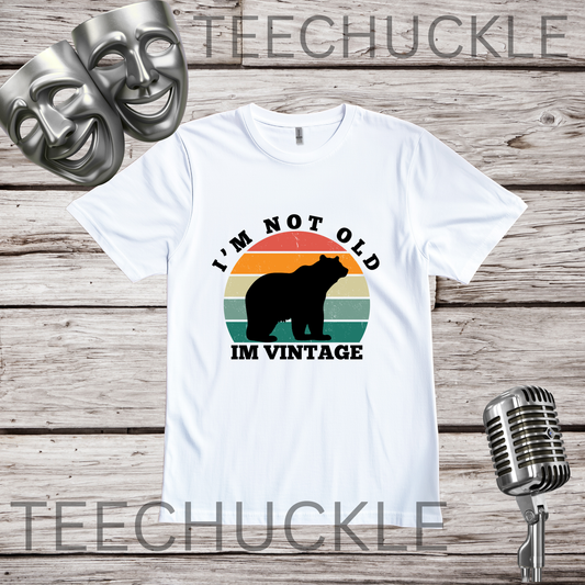 A white short-sleeve graphic T-shirt laid flat on a rustic wood background, with two silver theater masks and a vintage microphone as props. The shirt has a retro sunset-style bear graphic and the text 'I'M NOT OLD I'M VINTAGE.