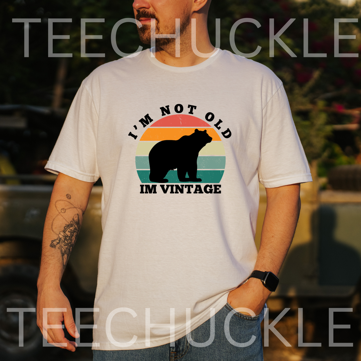 A man wearing a white graphic t-shirt with a retro sunset bear design and the text 'I'M NOT OLD IM VINTAGE'. The background is a blurred outdoor setting with a vehicle.