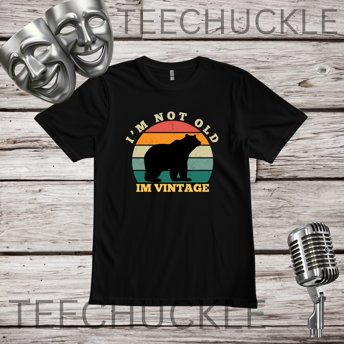 A black short-sleeve graphic T-shirt laid flat on a rustic wood plank background, with two silver theater masks, a vintage microphone. The shirt has a retro sunset-style bear graphic and the text “I'M NOT OLD IM VINTAGE.