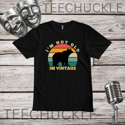 A black short-sleeve graphic T-shirt laid flat on a rustic wood plank background, with two silver theater masks, a vintage microphone. The shirt has a retro sunset-style bear graphic and the text “I'M NOT OLD IM VINTAGE.