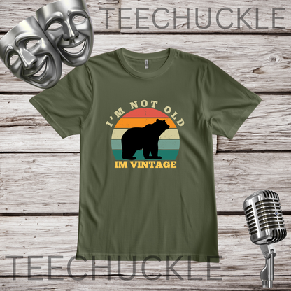 A green graphic t-shirt displayed on a rustic wooden background, featuring a retro-style bear silhouette and the text 'I'M NOT OLD IM VINTAGE'. The t-shirt is decorated with silver theater masks, a vintage microphone