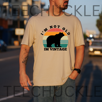 A man wearing a sand graphic t-shirt with a retro sunset bear design and the text 'I'M NOT OLD IM VINTAGE'. The background is a blurred street at sunset