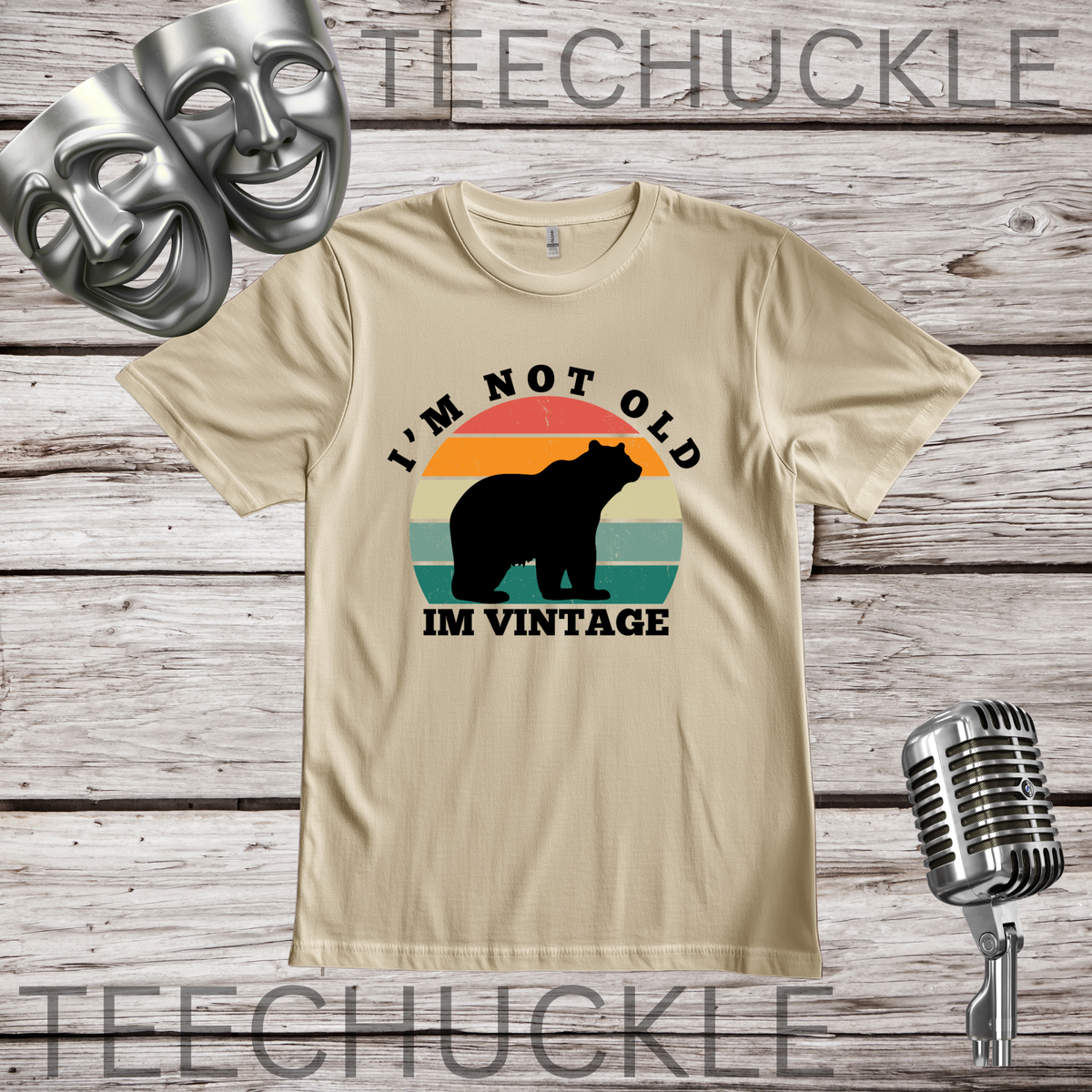 A sand graphic t-shirt displayed on a rustic wood background, featuring a retro design with a bear silhouette and the text 'I'M NOT OLD IM VINTAGE'. The shirt is styled with silver comedy and tragedy masks, a vintage microphone