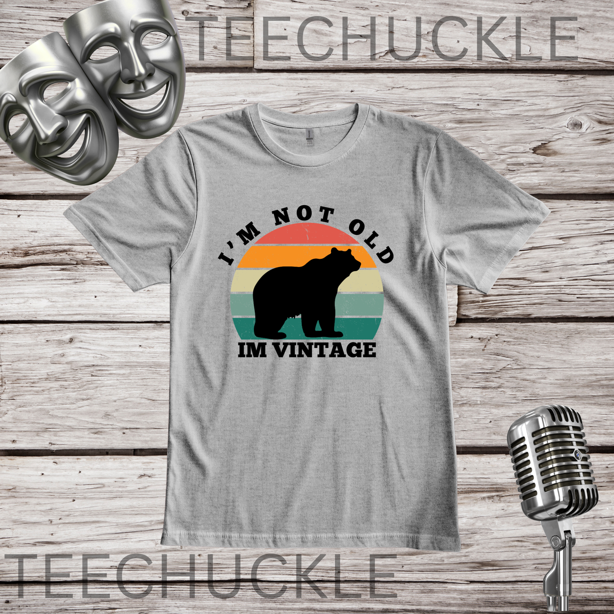 A gray crewneck t-shirt with a retro graphic of a bear in front of a sunset and the text 'I'M NOT OLD IM VINTAGE'. The shirt is displayed on a rustic wood background with a vintage microphone, comedy masks