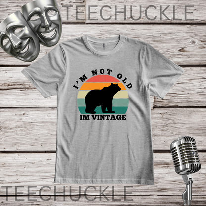 A gray crewneck t-shirt with a retro graphic of a bear in front of a sunset and the text 'I'M NOT OLD IM VINTAGE'. The shirt is displayed on a rustic wood background with a vintage microphone, comedy masks