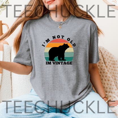 A woman wearing a gray graphic t-shirt with a retro sunset design and a black bear silhouette. The text reads 'I'M NOT OLD' above the sunset and 'IM VINTAGE' below.