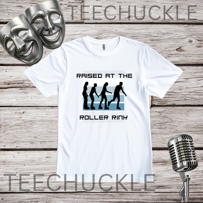 A white graphic t-shirt displayed on a rustic wood background, featuring a black and blue roller rink design with the text 'RAISED AT THE ROLLER RINK'. Two silver comedy masks and a vintage microphone are styled as prop.
