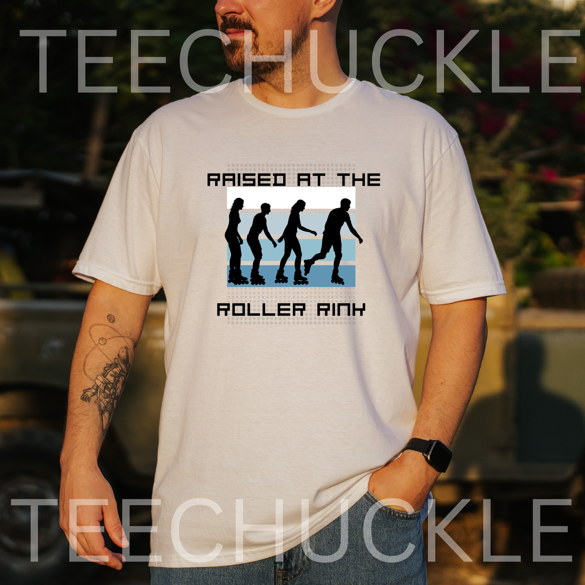 A man wearing a white graphic t-shirt with a black and blue roller rink design that reads 'RAISED AT THE ROLLER RINK'. The background is a blurred outdoor setting.