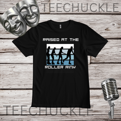 A black short-sleeve graphic T-shirt laid flat on a rustic wood plank background, with two silver theater masks and a vintage microphone as props. The shirt has a white and blue roller rink graphic with silhouettes and the text 'RAISED AT THE ROLLER RINK.