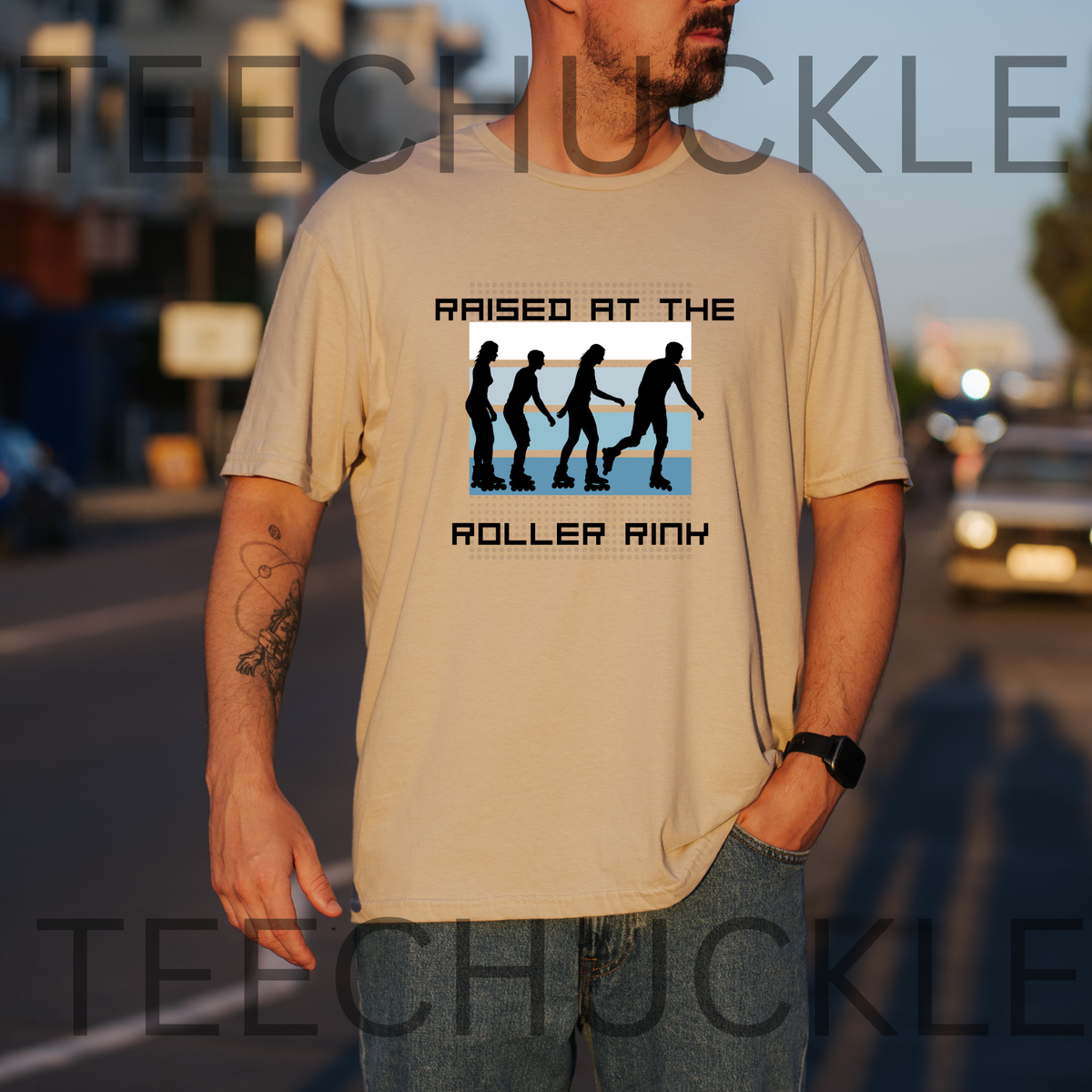 A man wearing a beige graphic t-shirt with a 'Raised at the Roller Rink' design, standing outdoors on a street at sunset. The t-shirt features a black and blue graphic of silhouettes walking in a row.