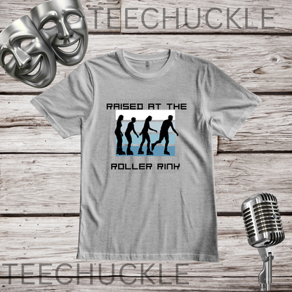 A gray graphic t-shirt displayed on a rustic wood background, featuring a black and blue graphic of silhouettes in roller skaters with the text 'RAISED AT THE ROLLER RINX'. The image includes decorative props like a silver microphone and theatrical masks