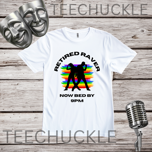 A white graphic t-shirt displayed flat on a rustic wood background, featuring a black silhouette of a dancing couple over a rainbow stripe pattern with the text 'RETIRED RAVER NOW BED BY 9PM'. Two silver comedy masks and a vintage microphone are styled as props.