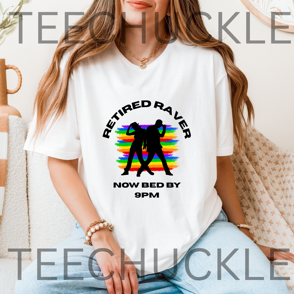 A woman wearing a white graphic t-shirt with a black silhouette of two people dancing in front of a rainbow sunset, with the text 'RETIRED RAVER NOW BED BY 9PM'.