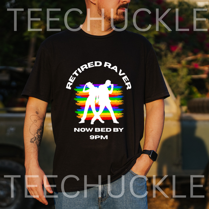 A man wearing a black graphic t-shirt with a colorful rainbow design and white text that reads 'RETIRED RAVER NOW BED BY 9PM'. The background is an outdoor setting with a blurred vehicle and foliage.
