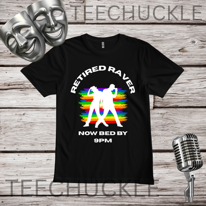 A black short-sleeve graphic T-shirt laid flat on a rustic wood plank background, with two silver theater masks, a vintage microphone. The shirt graphic shows rainbow stripes with a white silhoutte of 2 dancers with the words “RETIRED RAVERS NOW BED BY 9PM” 