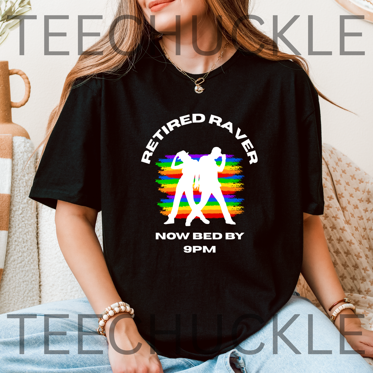 A woman wearing a black graphic t-shirt with a white text design that reads 'RETIRED RAVER NOW BED BY 9PM' over a colorful rainbow-striped background with two white silhouettes. 
