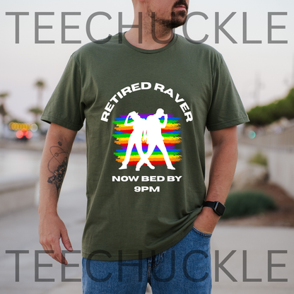 A man wearing a green graphic t-shirt with a 'Retired Raver Now Bed By 9PM' design featuring a rainbow and dancing figures, set against a blurred outdoor background. 