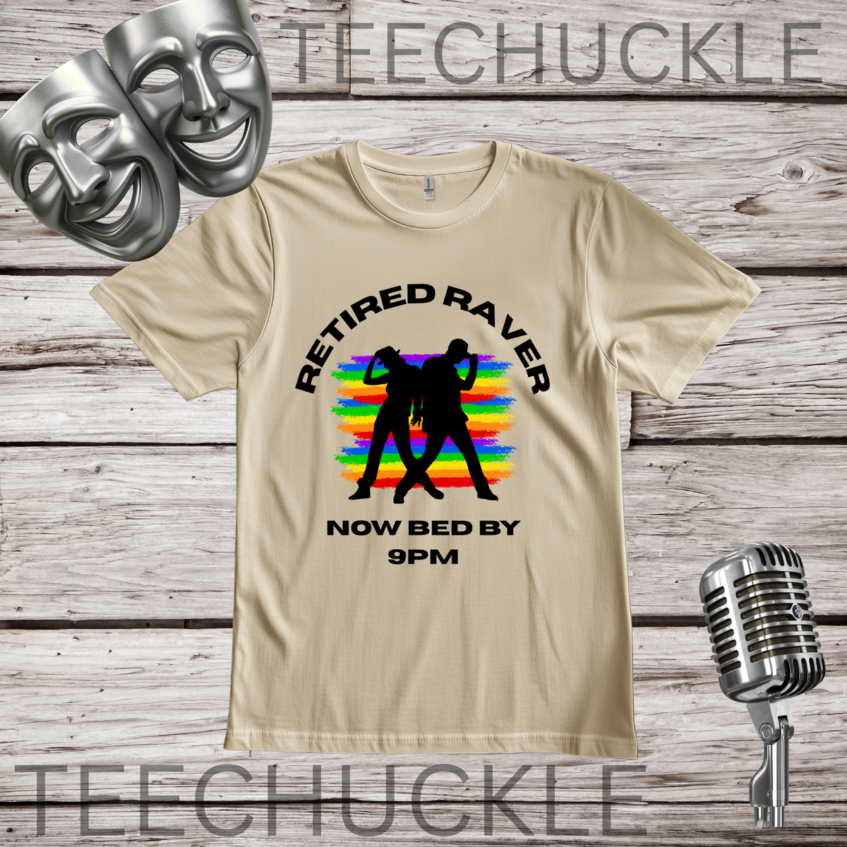 A sand graphic t-shirt displayed on a rustic wood background, featuring a black text design that reads 'RETIRED RAVER NOW BED BY 9PM' with a black silhouette of two people against a rainbow sunset. The shirt is decorated with silver theater masks, a vintage microphone