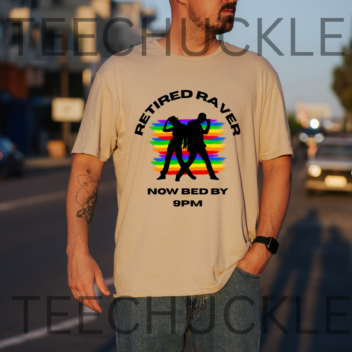 A man wearing a sand graphic t-shirt with a black 'Retired Raver Now Bed by 9PM' design featuring a rainbow stripe background and black silhouettes, standing on a blurred street at sunset.