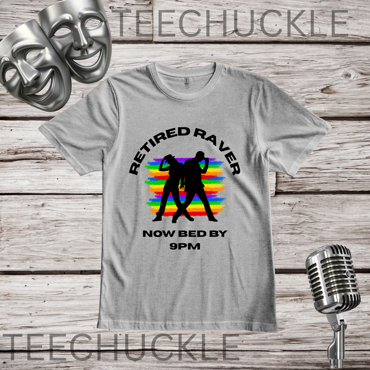 A gray graphic t-shirt displayed on a rustic wooden background, featuring a black silhouette of a couple on a rainbow sunset with the text 'RETIRED RAVER NOW BED BY 9PM'. The image includes decorative props like a vintage microphone, comedy and tragedy masks, and a silver microphone stand.